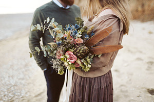 Newlyweds with rustic bouquet in hands stand on seashore. Bride and groom wear in country style clothes and hats.