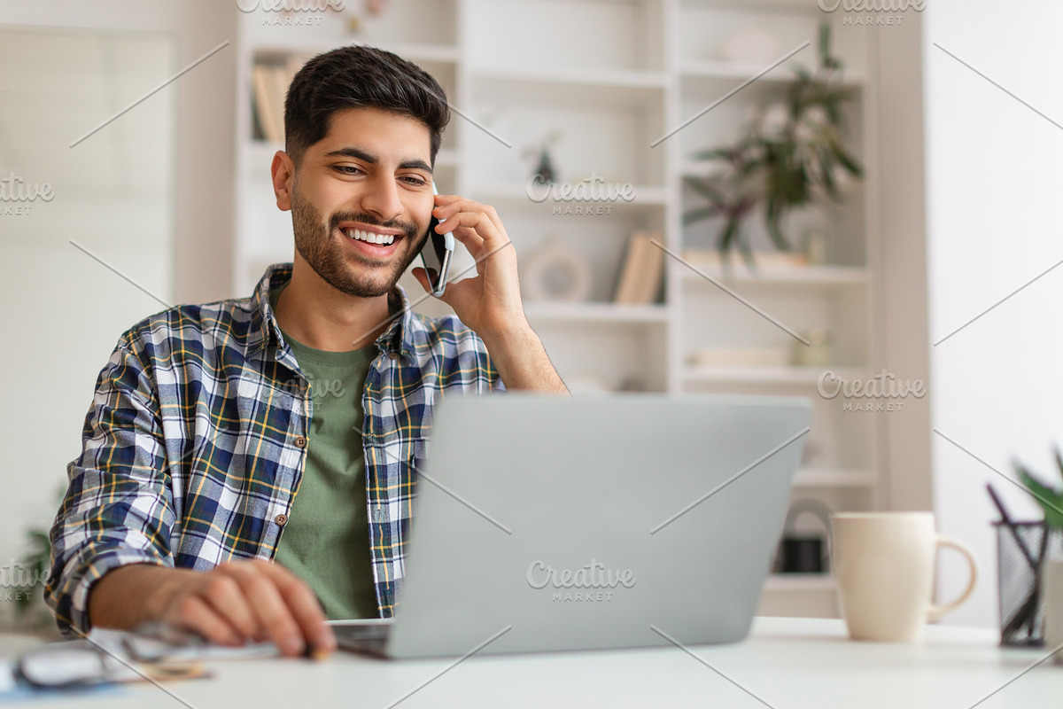Smiling Arab man working and talking on phone at home, a Photo by Prostock-Studio (Photo 2 of 2)