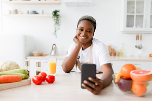 Cheerful millennial black woman in apron reads message, makes selfie on phone a, a Person Photo by Prostock-Studio