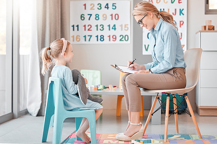 Full length caucasian child psychologist sitting with an adorable little girl in a clinic and using a clipboard for a consult. Cute girl talking to a mental health professional. Dyslexia and anxiety, a Photo by Peopleimages.com