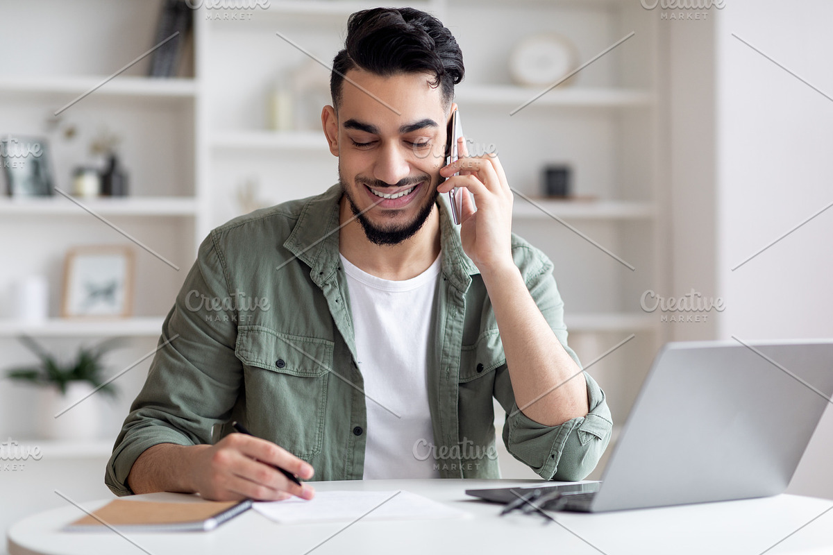 Handsome Arab Male Freelancer Talking On Cellphone And Taking Notes At Home, a Photo by Prostock-Studio (Photo 2 of 2)