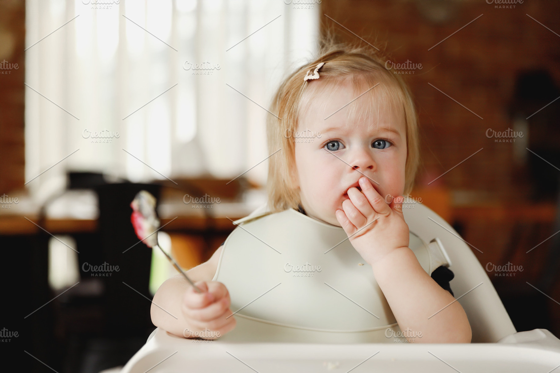 Infant girl or toddler sitting on high chair. Eating food by herself using spoo, a Person Photo by Lenor4ik