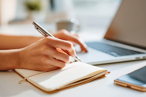 Person writing in a notebook beside a laptop, a Business Photo by AlyaAnd