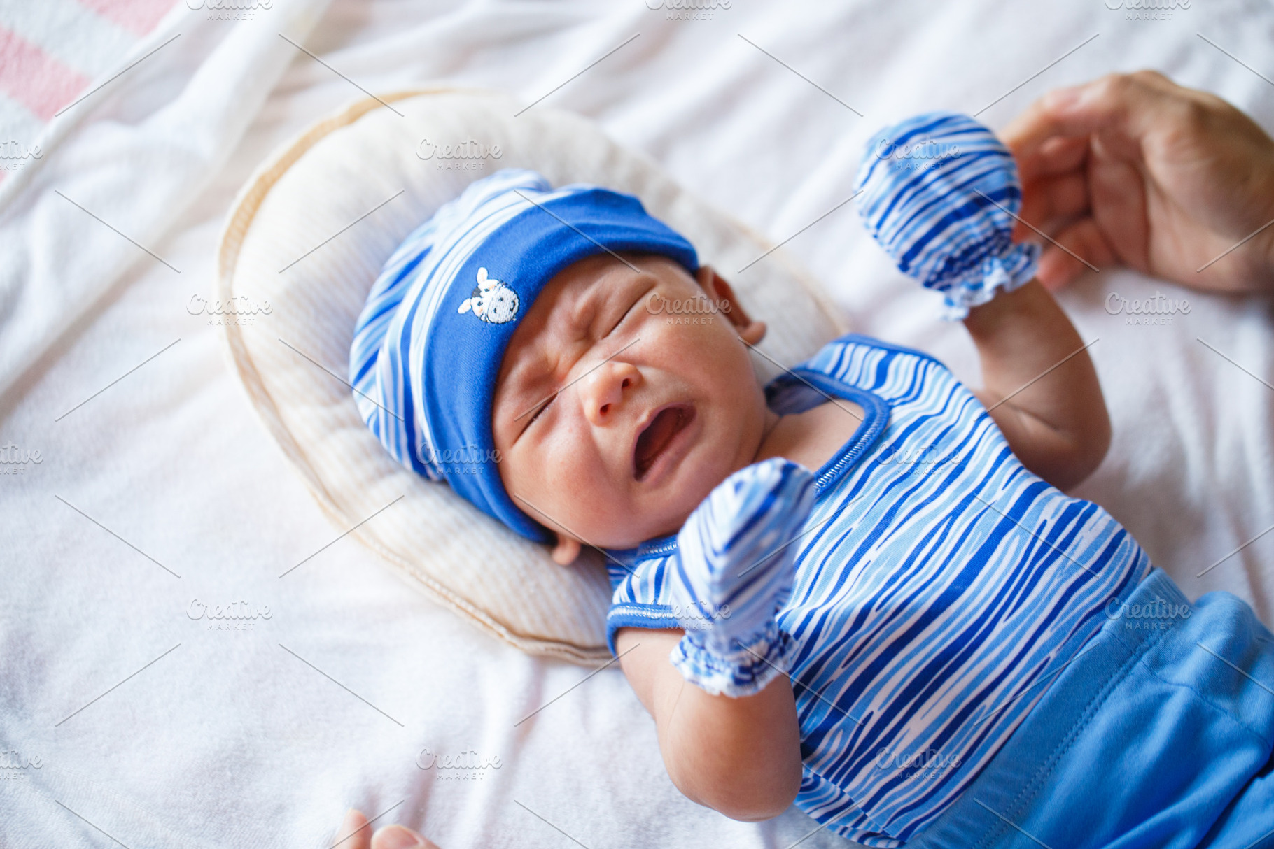 Crying baby in blue outfit with hat and mittens on soft bedding, a Person Photo by a454