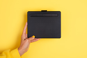 Hand holding black digital drawing tablet against vibrant yellow, a Technology Photo by Ales Munt
