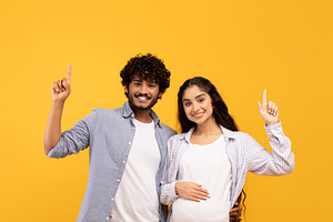 Excited indian pregnant lady and her husband pointing up at free space, showing, a Person Photo by Prostock-Studio
