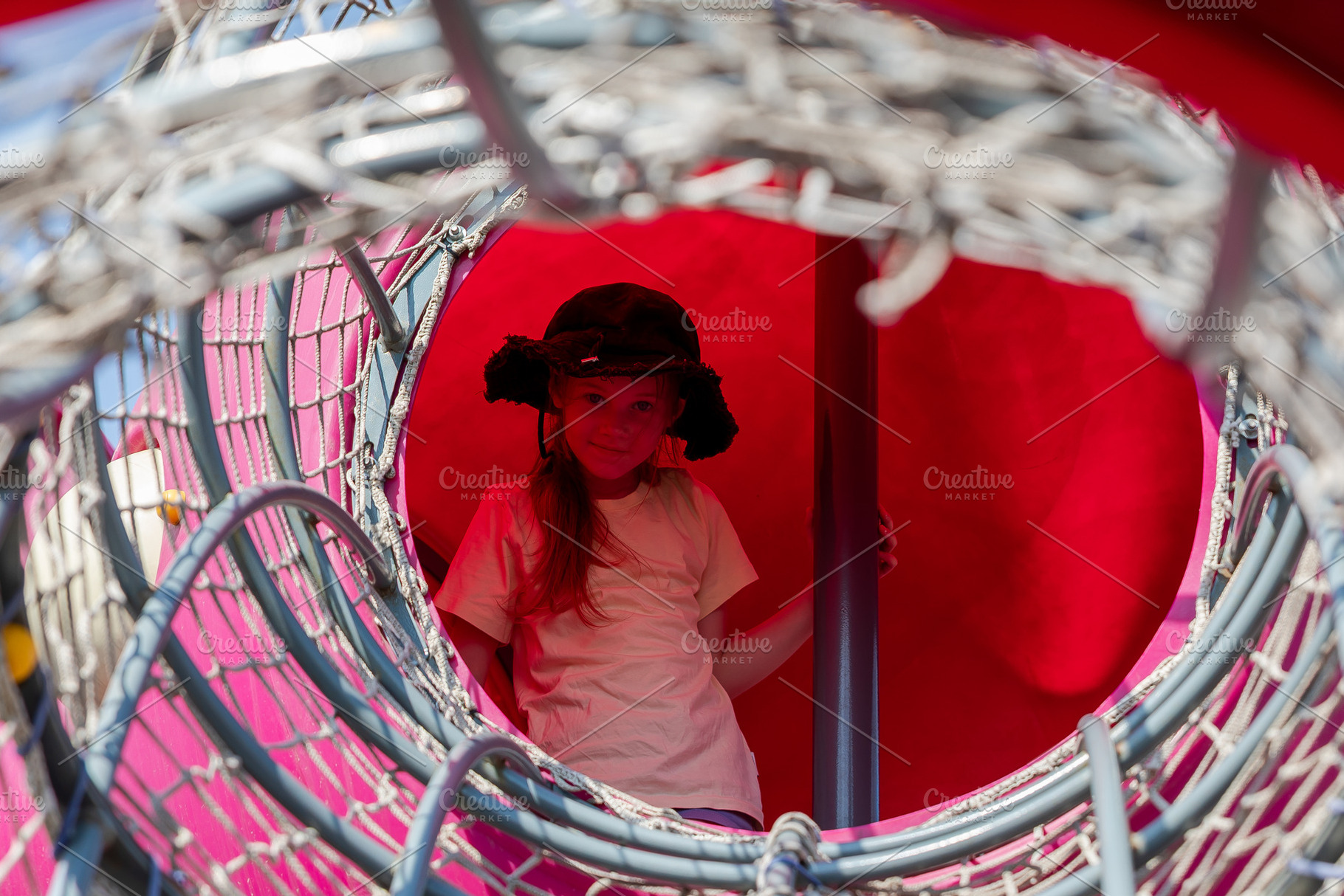 Smiling young girl in a sun hat stands inside a colorful red playground tunnel,, a Person Photo by Ales Munt