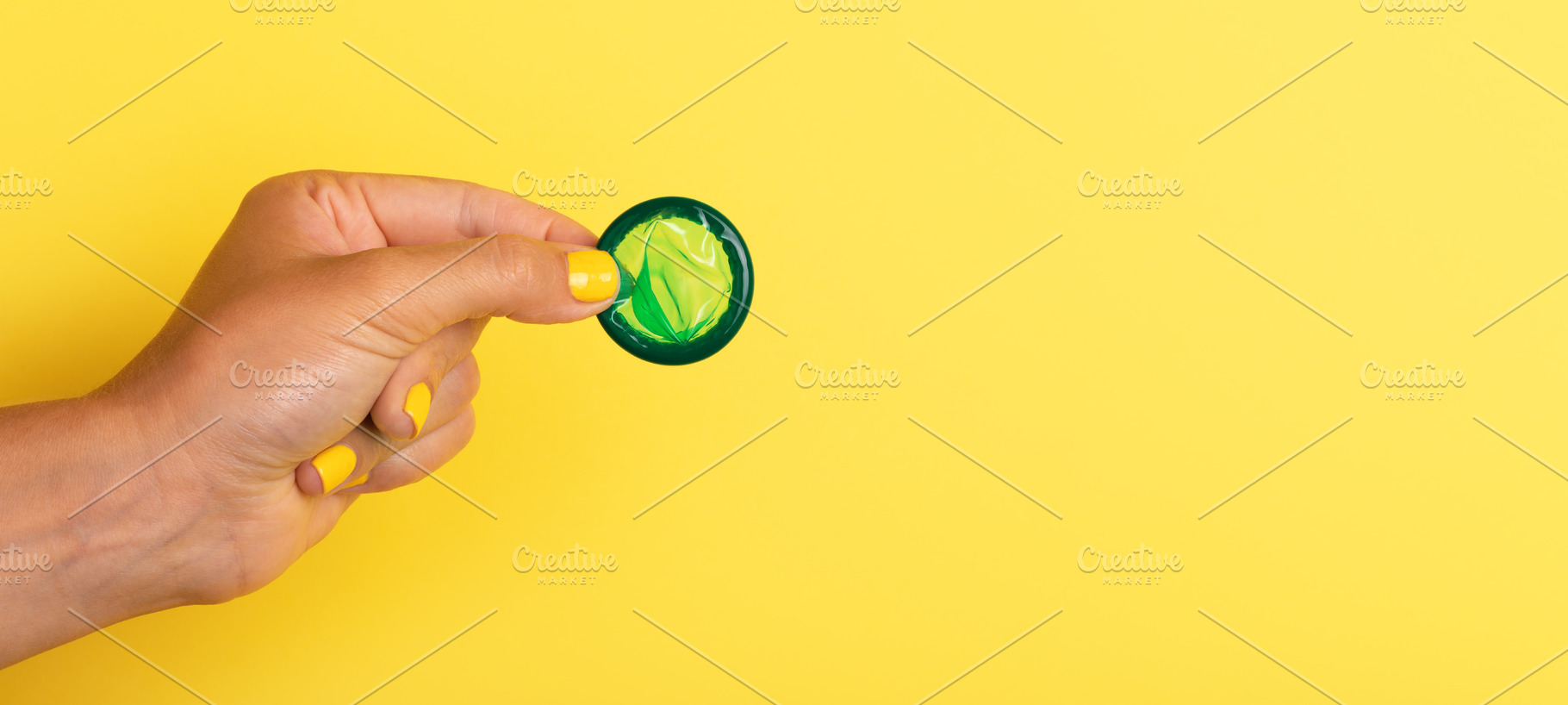Hand with yellow nails holding a green rolled condom against a, a Health & Medical Photo by Ales Munt