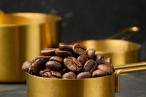 Roasted coffee beans are heaped into a golden metal measuring cu, a Food & Drink Photo by ndanko