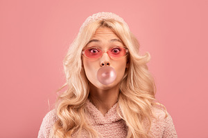 Joyful blonde lady chewing gum and blowing bubble, looking at camera on pink st, a Photo by Prostock-Studio