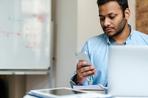 Middle eastern unshaven man using mobile phone while working with laptop