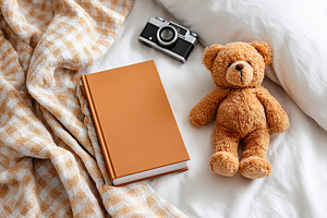 Blank hardcover book on bed alongside teddy bear and vintage camera in cozy set, a Photo by AlyaAnd