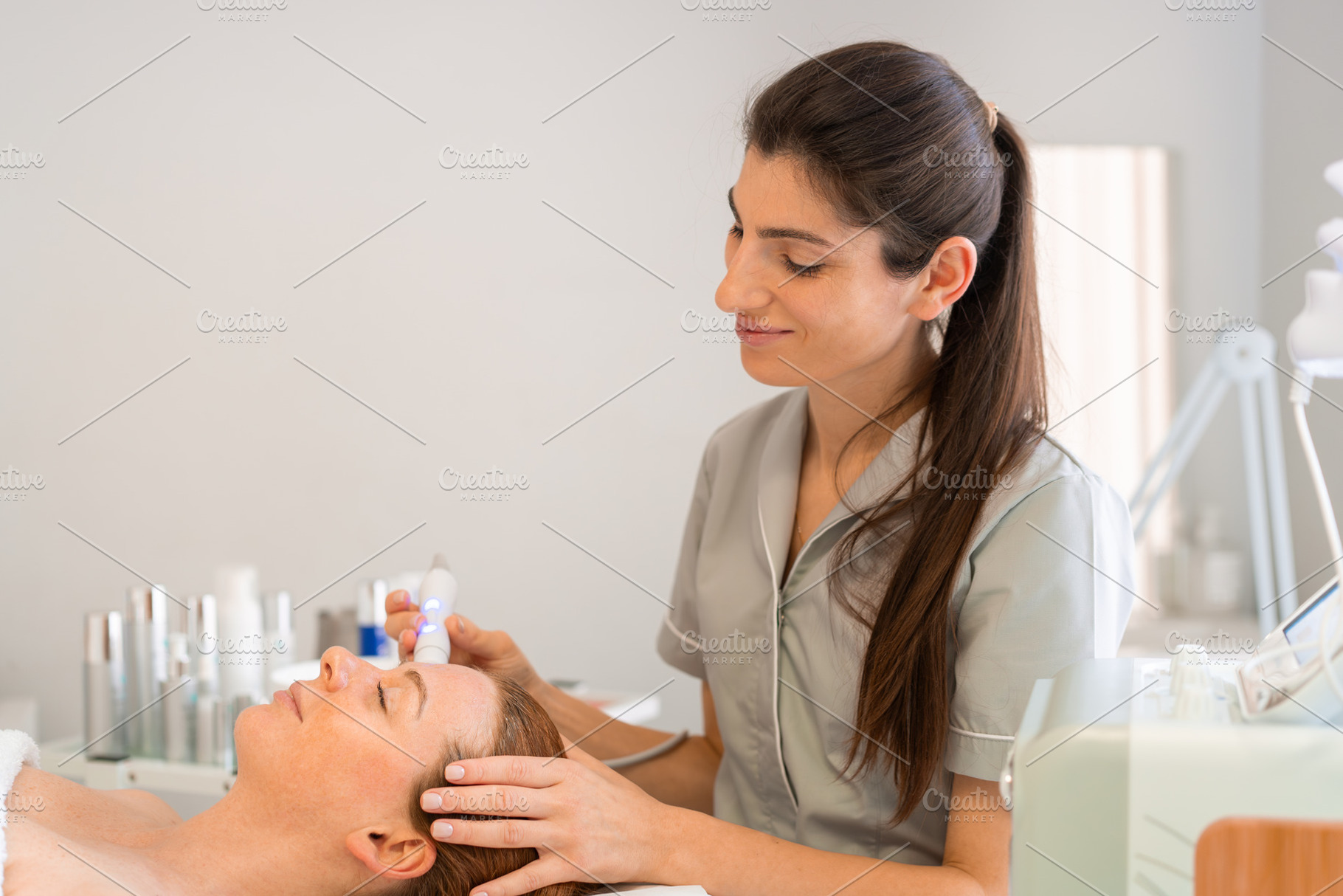 Smiling female cosmetologist applying iontophoresis on client's face, a Person Photo by andreonegin market
