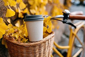 Coffee cup mockup placed inside a bicycle basket surrounded by autumn leaves, a Photo by AlyaAnd