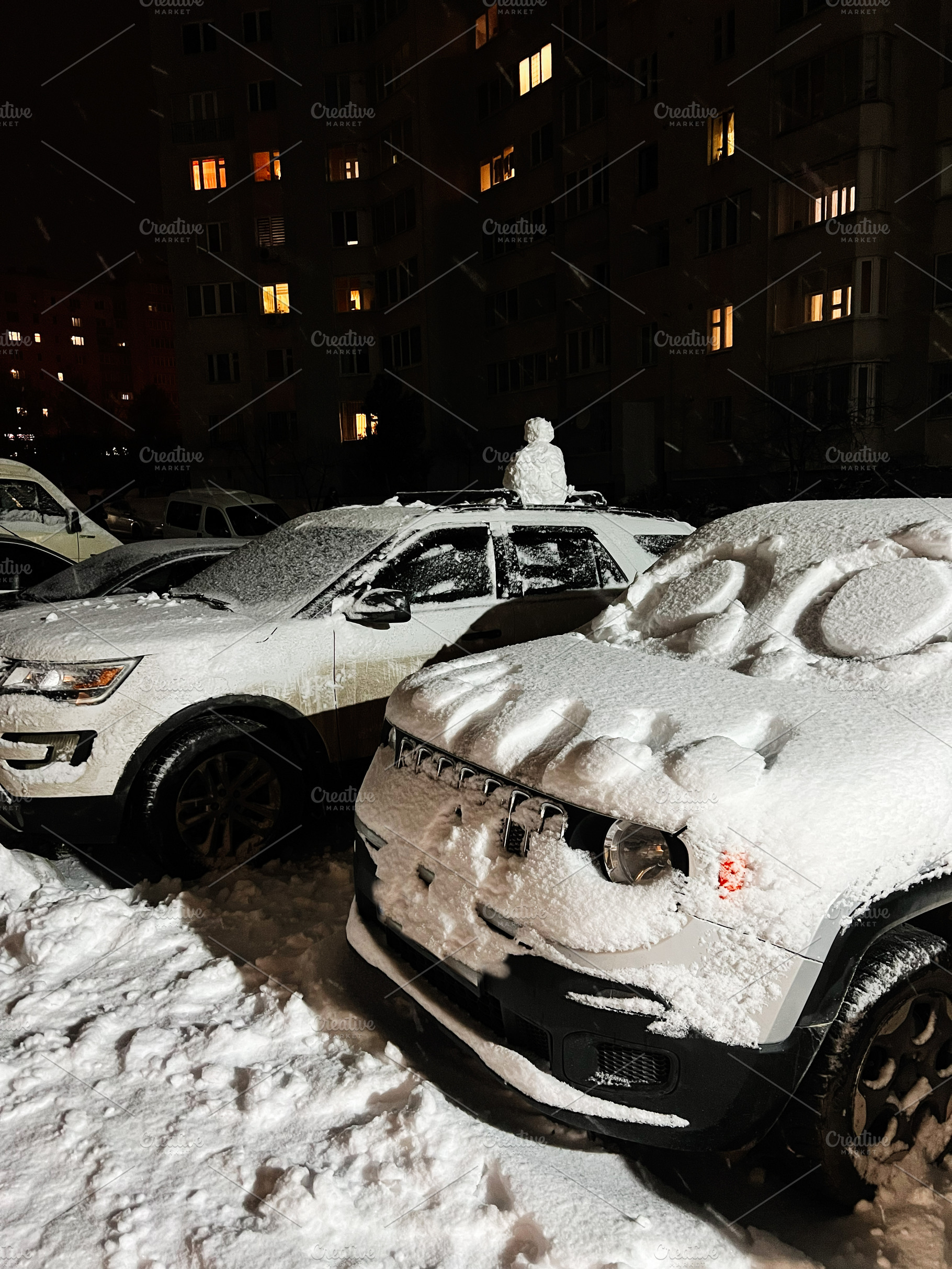 Snowy night in a city parking lot with snow covered cars and glowing apartment, a Photo by AS photostudio