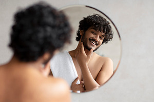 Young indian man looking in mirror in bathroom and touching beard, handsome guy, a Person Photo by Prostock-Studio