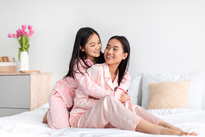 Smiling japanese teenage girl in pajama hugs young woman on bed and have fun in, a Photo by Prostock-Studio