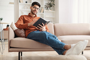 Smiling Arab man in glasses reading paper book, a Photo by Prostock-Studio