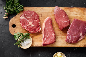 Different cuts of raw beef on a wooden cutting board with herbs and butter in a