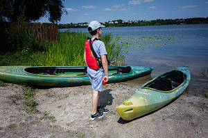 Boy standing on riverbank with kayaks ready for paddling under blue skies, a Photo by AS photostudio