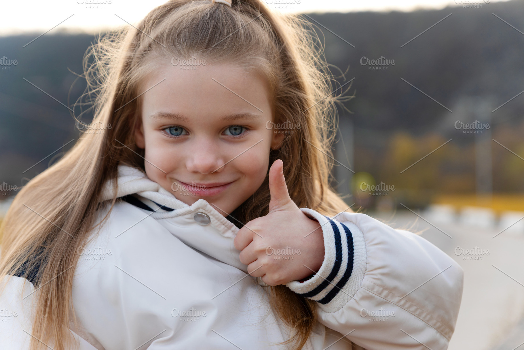 Cheerful young girl in a white jacket showing thumbs up outdoors, a Person Photo by Ales Munt