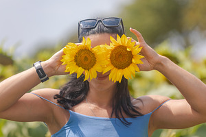 Smiling woman in blue top playfully covers eyes with sunflowers in a sunny outd, a Person Photo by Ales Munt