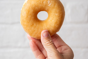 Hand Holding Glazed Donut Against White Brick Background, a Food & Drink Photo by etorres69