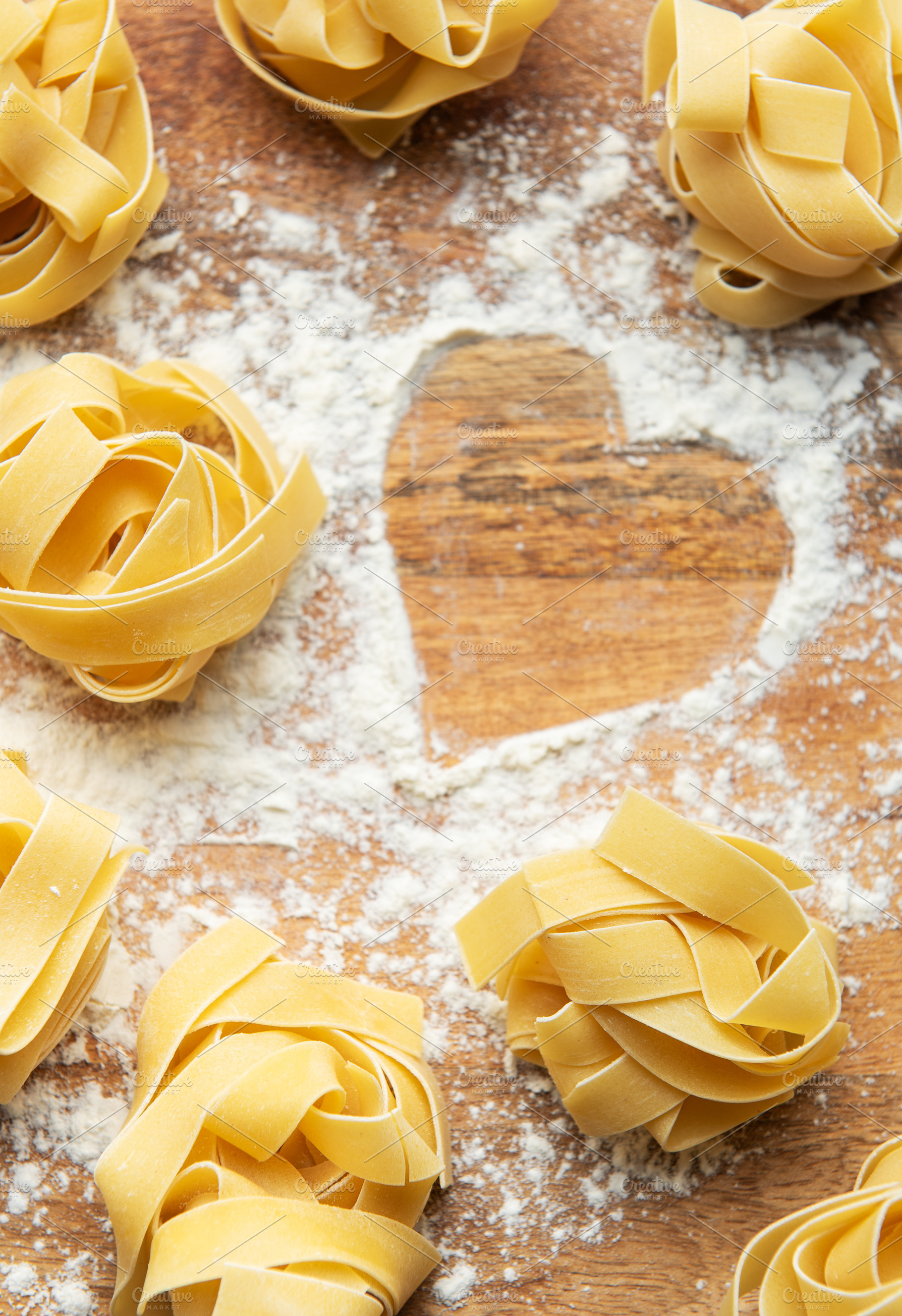 Uncooked tagliatelle pasta nests with heart of flour on wooden b, a Food & Drink Photo by Olena Rudo