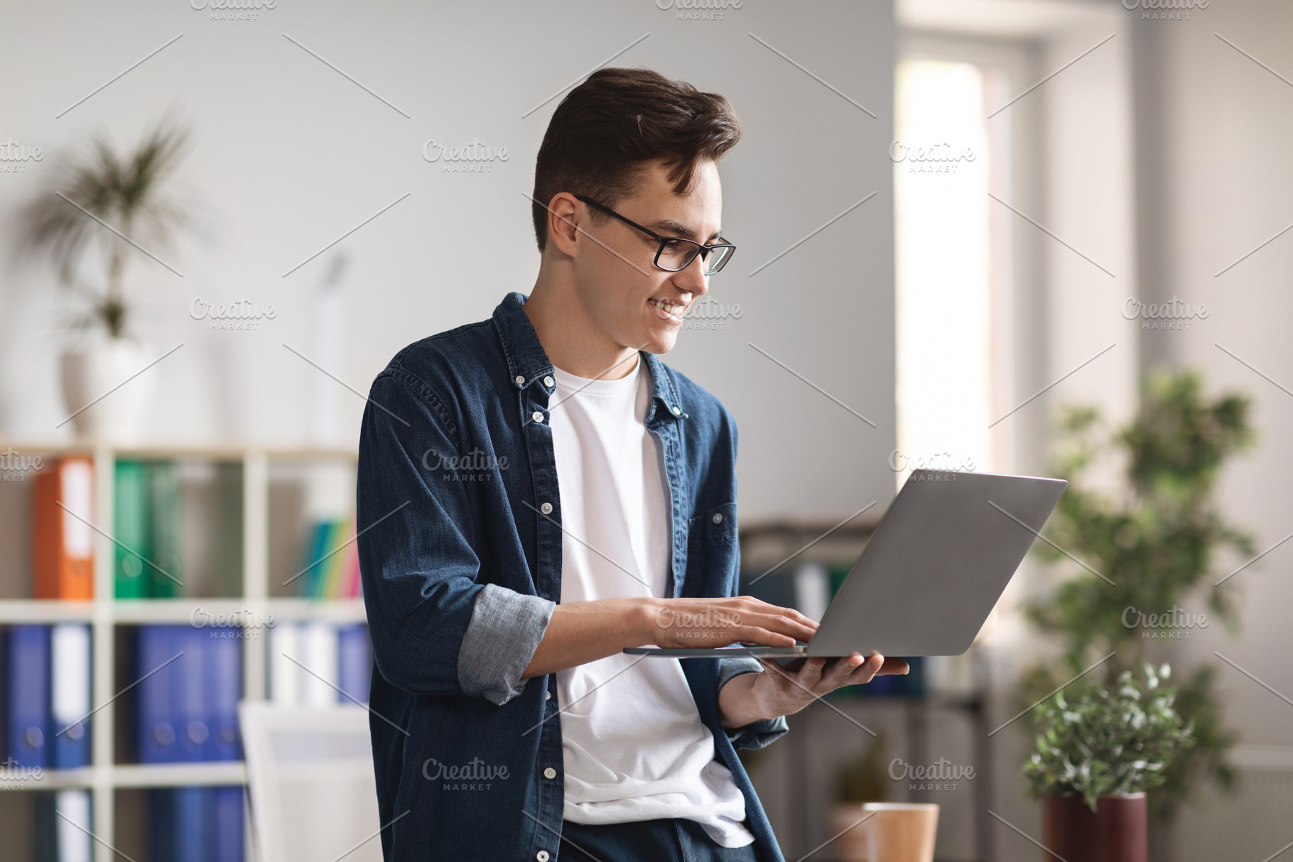 Smiling Millennial Man Using Laptop While Standing Near Desk In Modern Office, a Photo by Prostock-Studio