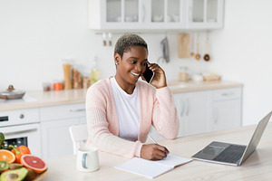 Smiling millennial pretty black lady manager speaks by phone and looks at lapto, a Person Photo by Prostock-Studio