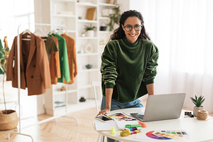 Happy Clothing Designer Lady Using Laptop Smiling Standing In Showroom, a Photo by Prostock-Studio