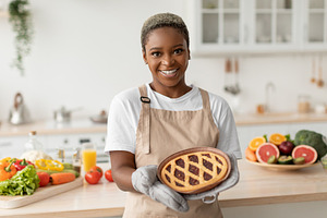 Smiling pretty millennial african american woman in apron showing tasty pie in, a Person Photo by Prostock-Studio