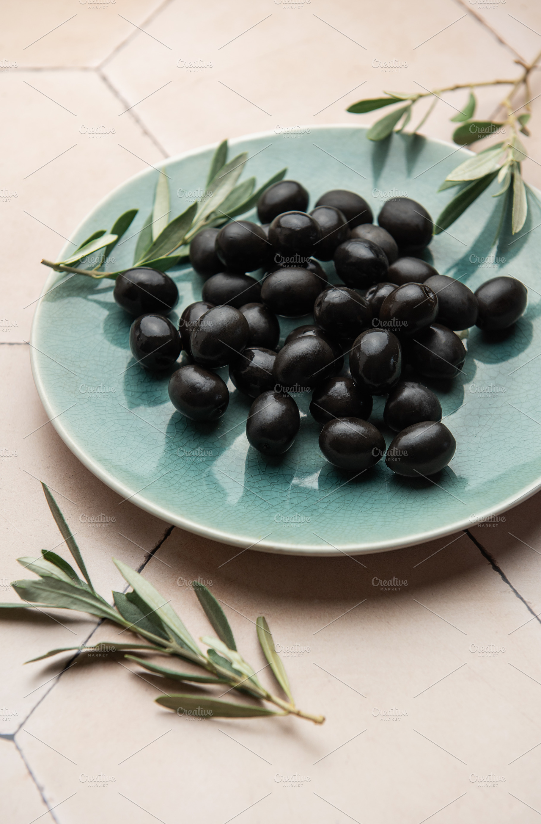 Black olives served on ceramic plate with olive branches on tile, a Food & Drink Photo by Olena Rudo
