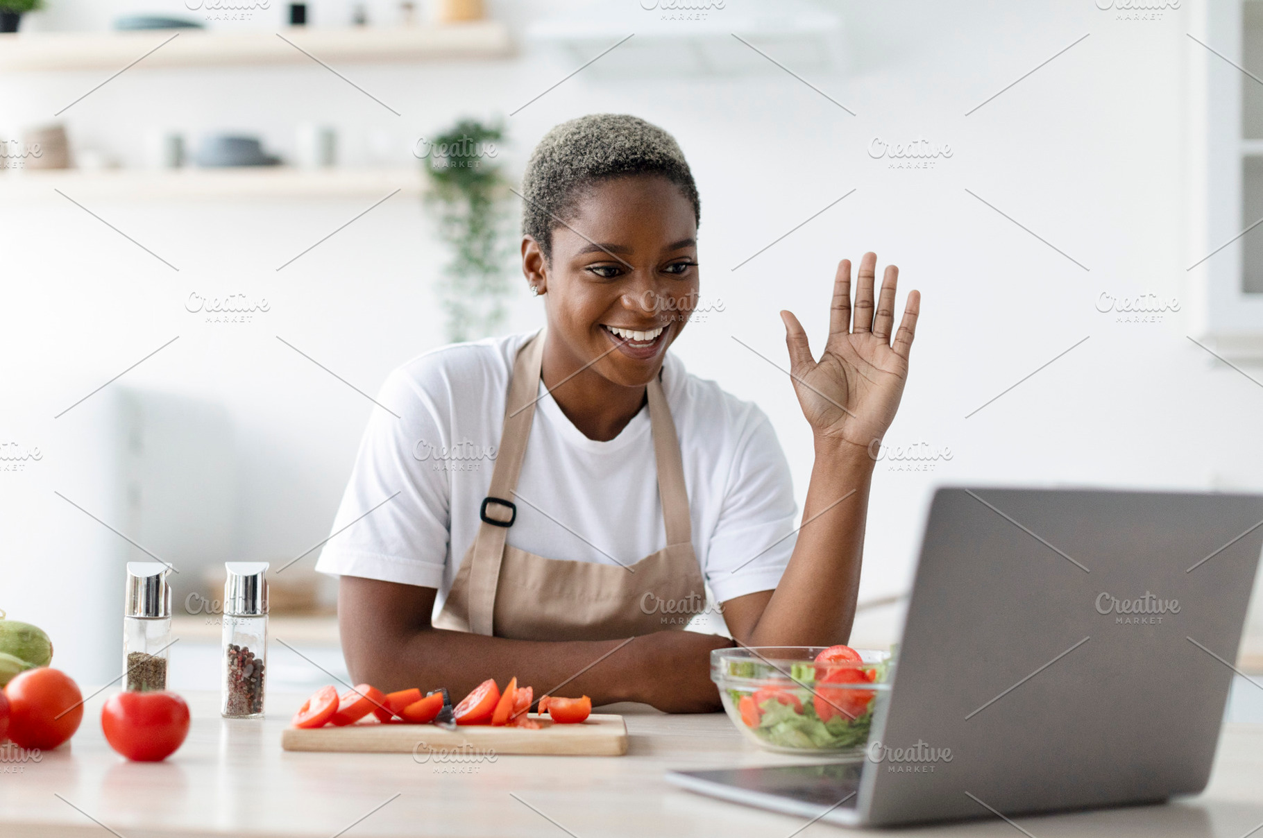 Glad millennial african american female in apron waving hand at laptop in kitch, a Person Photo by Prostock-Studio