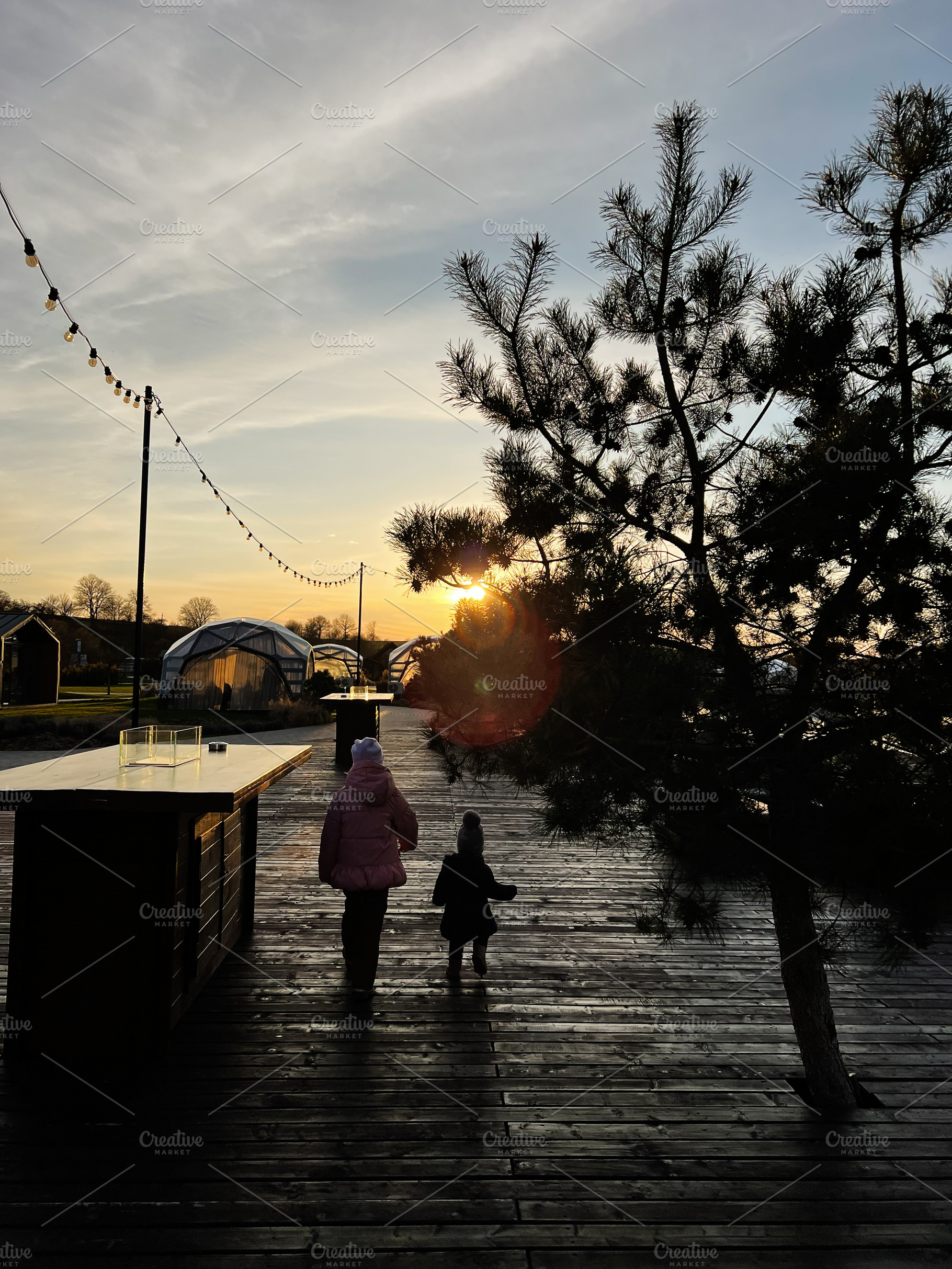 Two people walking on a sunset-lit wooden path near outdoor structures, a Photo by AS photostudio