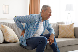 Mature man with side back pain sitting on couch at home, a Photo by Prostock-Studio
