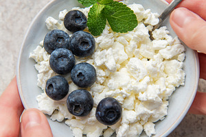 Hands holding a bowl of cottage cheese with blueberries and mint, a Food & Drink Photo by Olena Rudo