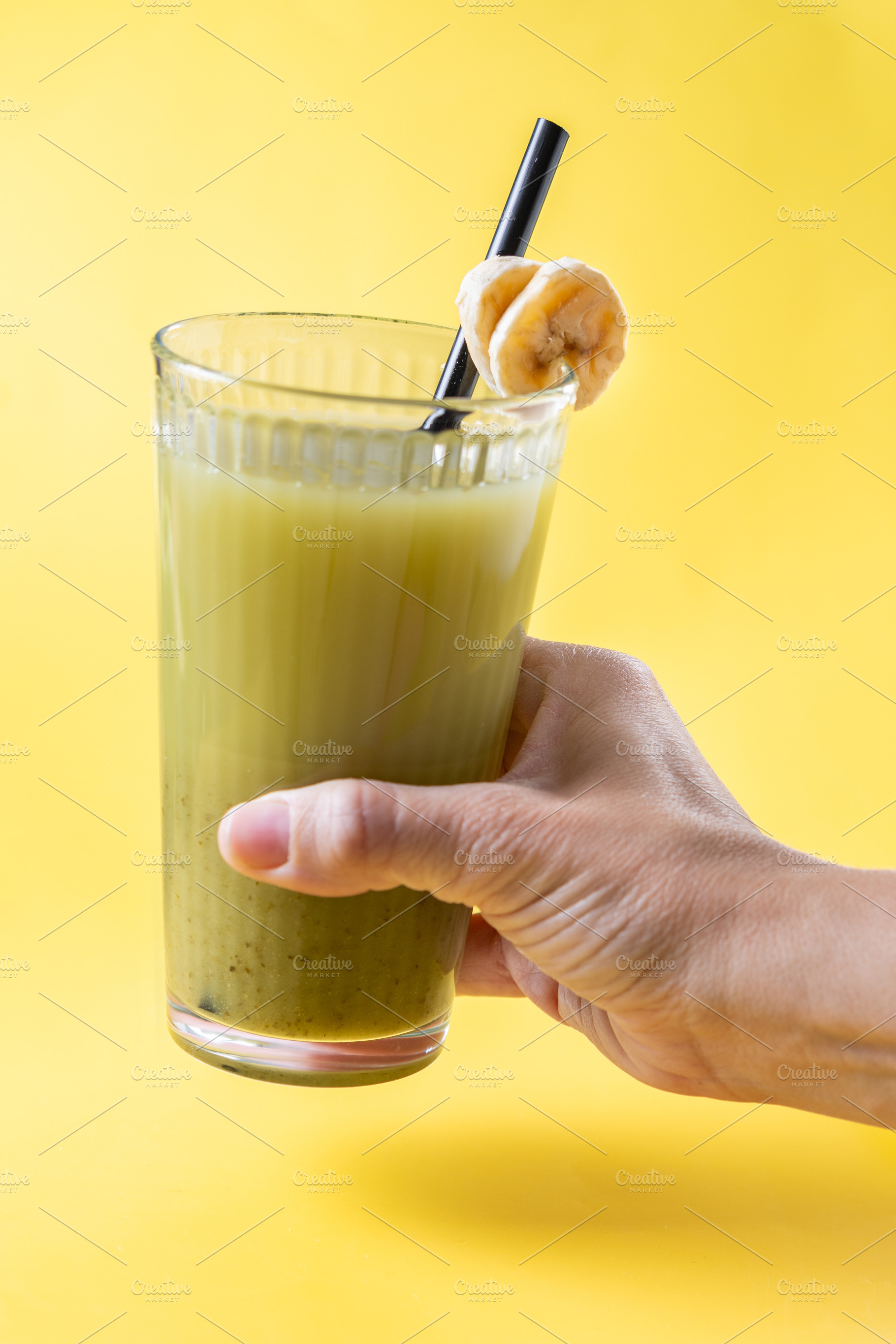 Hand holding a green smoothie with banana on a yellow background, a Food & Drink Photo by etorres69