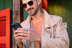 Smiling man in beige trench coat using smartphone outside green house, a Person Photo by andreonegin market
