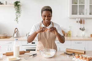 Cheerful millennial african american lady in apron breaks egg preparing dough i, a Photo by Prostock-Studio