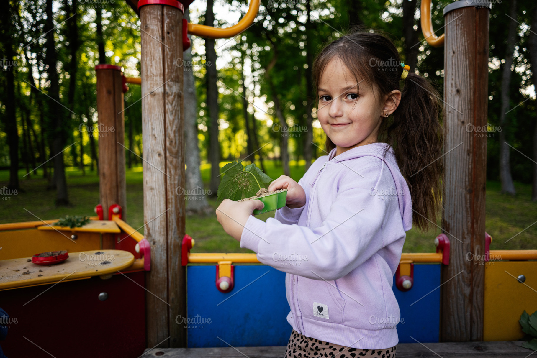 Smiling girl holding green leaves at a colorful playground in a forested park, a Person Photo by AS photostudio