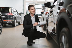 Happy young car salesman sitting near new auto, checking it, taking notes at au, a Person Photo by Prostock-Studio