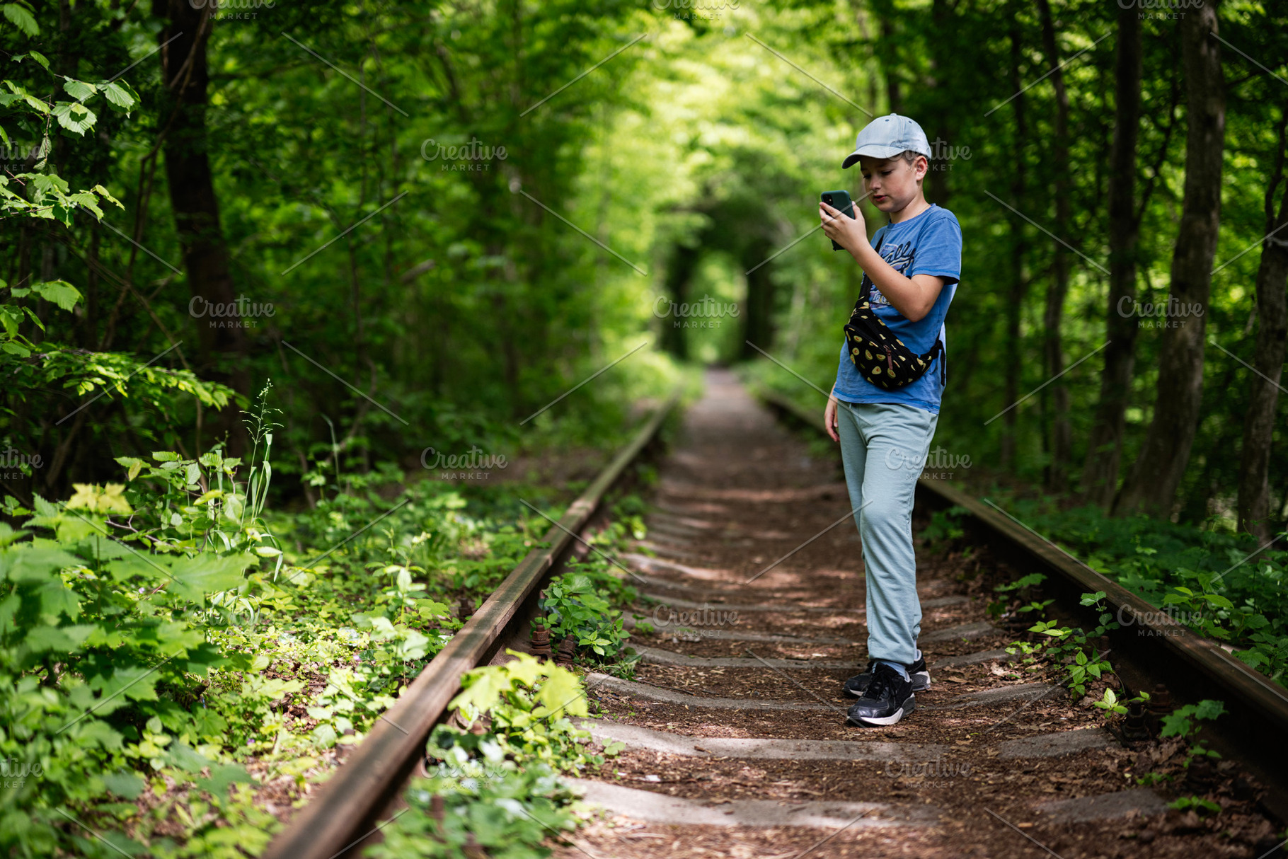 Boy walking on railway tracks and using mobile phone in green forest, a Photo by AS photostudio
