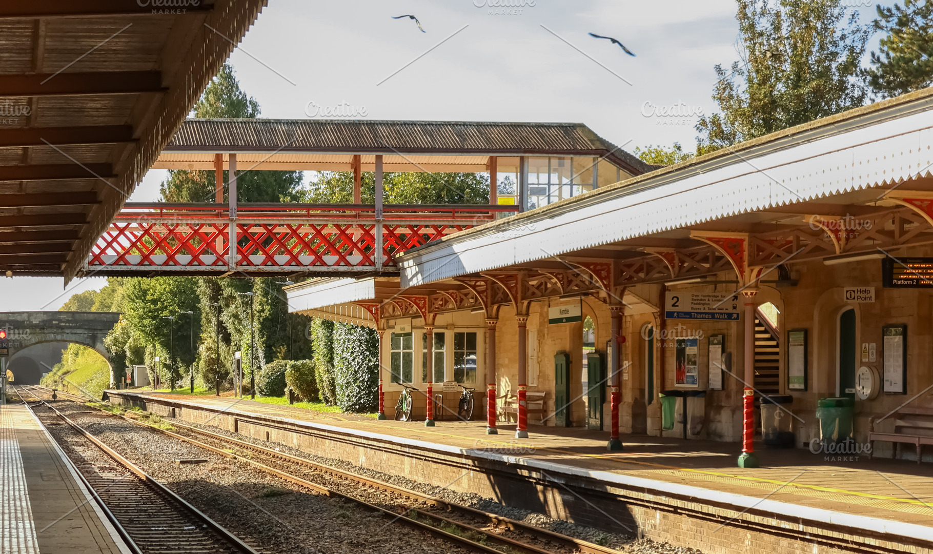 Vintage train station platform and red footbridge with tracks and signals. Kemb, a Photo by Studio ANNA