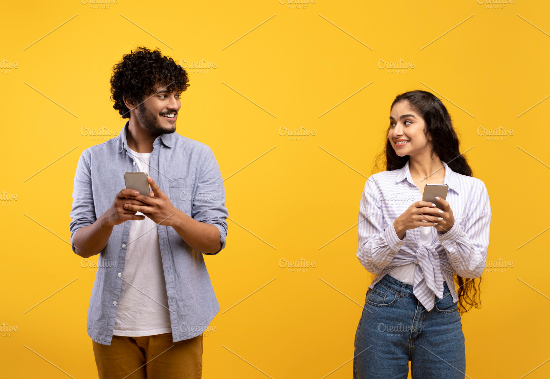 Cheerful young indian man and woman in casual outfits holding smartphones, look, a Photo by Prostock-Studio