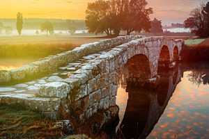 Ancient stone bridge with arched spans crosses calm river at sun, a Nature Photo by José Manuel Gelpi