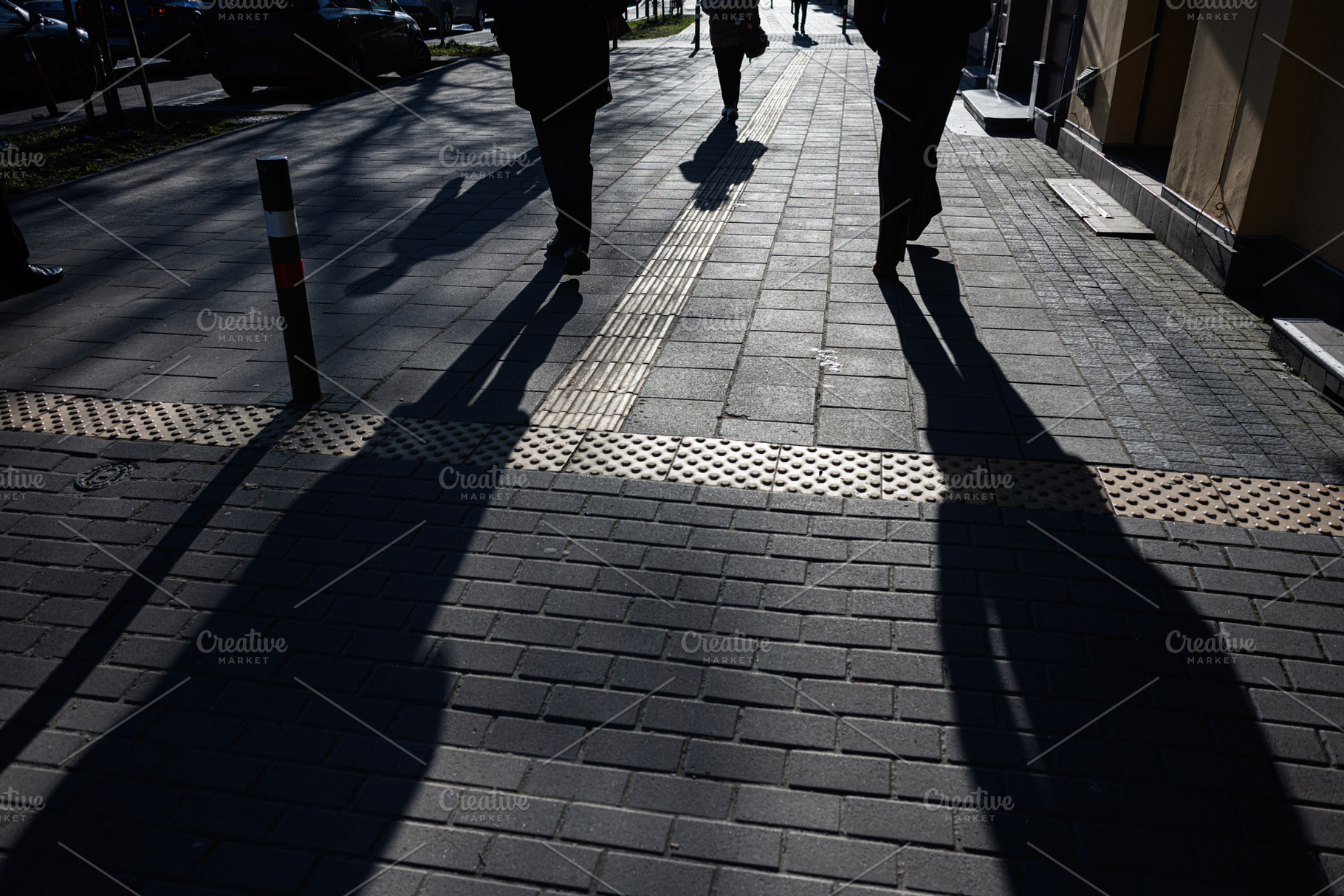 Shadows of pedestrians on sunlit urban sidewalk with textured tactile paving, a Photo by AS photostudio