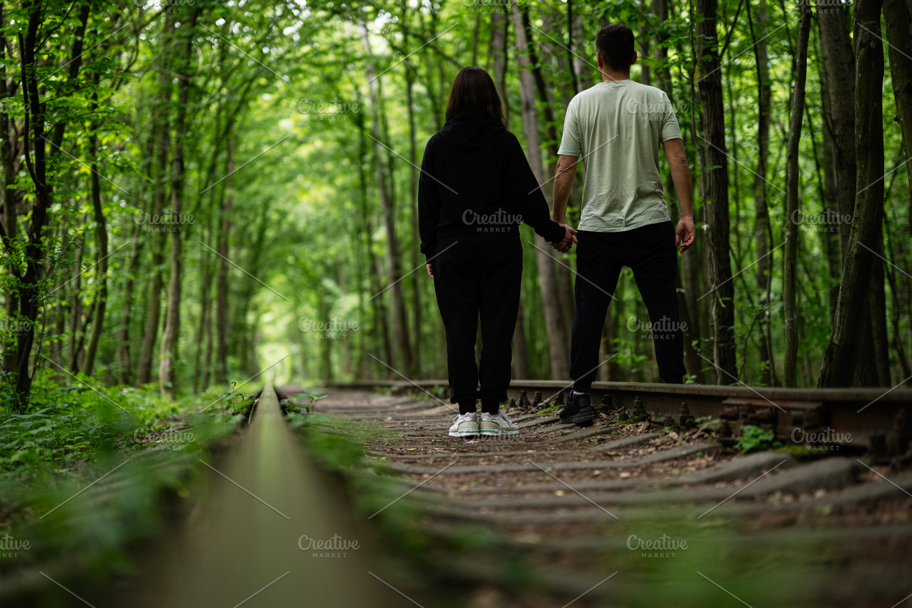 Couple holding hands walking along a forest path between trees on train tracks, a Photo by AS photostudio