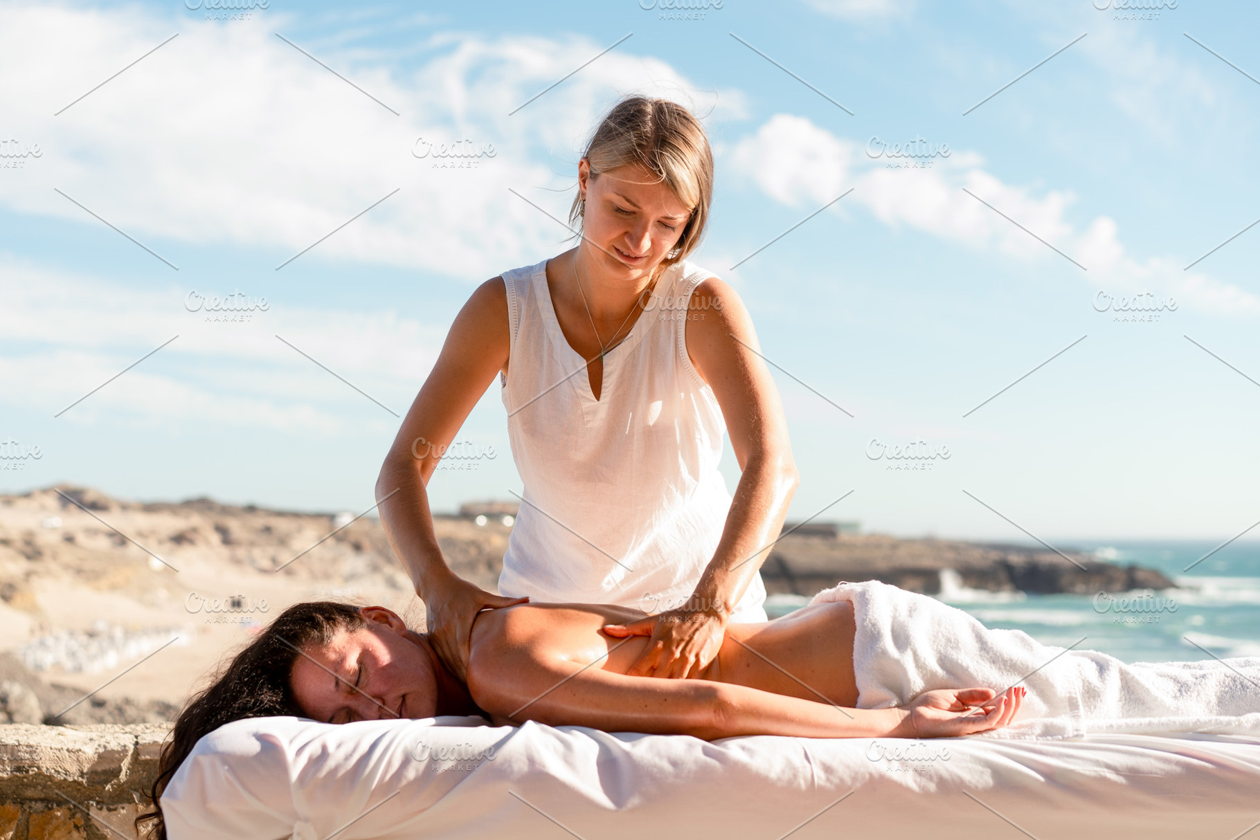 Therapist massaging woman's back on table at beach during sunny day, a Photo by andreonegin market