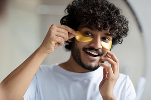 Skin care to reduce eye bags. Cheerful indian man applying gold anti-fatigue pa, a Person Photo by Prostock-Studio